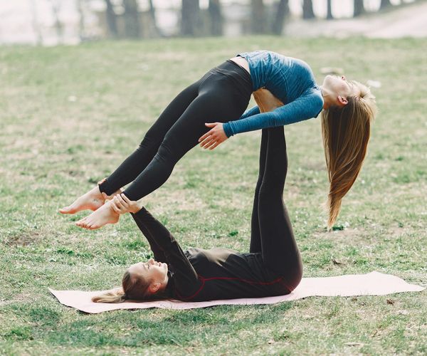 Two friends laughing while attempting a simple partner yoga pose outdoors.